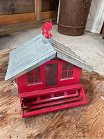 Metal bird house with red body, silver roof, and red decorative bird on top, placed on wooden surface.