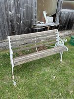 Outdoor park bench photographed on grass next to a weathered wooden structure, showing full view of bench with missing wooden slats and weathered cast iron armrests.
