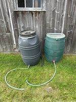 Two rain barrel buckets, one black larger barrel with removable top and one smaller green barrel, both connected with hoses, placed outside against wooden fence.