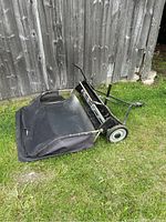 Full view of tow-behind sweeper and attached grass catcher bag leaning against wood fence