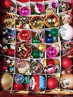 Box with divided sections containing a variety of shiny Christmas bulbs in multiple colors including red, gold, green, pink, blue, white, and multi-color hand-painted ones.