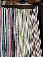 Top-down view of a box stacked with a large number of assorted vintage vinyl records in sleeves.