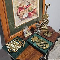 Overall lot view showing brass pineapple doorstop, two pineapple trivets/hotplates, brass and silver candlestick holders on wooden surface next to framed artwork