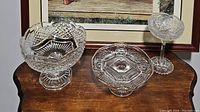 Three clear crystal pedestal dishes arranged on a wooden surface under a framed picture.