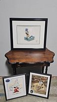 Three framed Disney sericel art pieces displayed on and in front of a wooden table.