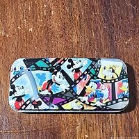 Colorful Disney character filmstrip design tin featuring Mickey Mouse, Donald Duck, and Goofy with vibrant colors on a wooden surface.