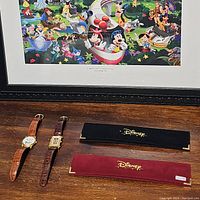 Overview photo showing two Disney watches with leather bands and two Disney branded watch boxes placed on a wooden surface with Disney character collage artwork in background.