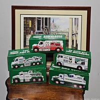 Seven boxed Hess toy vehicles stacked on a table against a framed picture, showing brand and toy types on packaging.