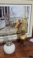 Lot shows two vintage oil lamps side by side in front of a framed painting. One lamp has a white round base and clear chimney. The other is tall amber colored with coin pattern glass.