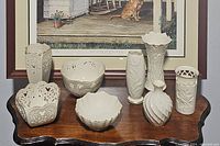 Group of seven white Lenox porcelain vases and bowls with varying embossed and pierced designs, displayed on a wooden table under a framed painting.