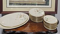 Stack of 12 dessert plates, 12 saucers, and 2 oval platters with leaf pattern on wooden table under framed picture