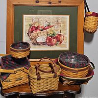 Photo showing six Longaberger baskets arranged on a wooden table with fabric liners and plastic protectors, variety of basket shapes including round and rectangular, one with leather handles, and background wall art featuring apples.