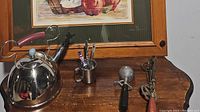 Photo showing the five kitchen items on a wooden table: the shiny Mickey Mouse themed kettle, ice cream scoop, egg beater, tin cup with forks, and cheese spreader.