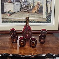 Full shot of the red Murano decanter set with six cups arranged around it, showing the gold trim and design details.
