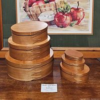 Six Briarwood nesting oval boxes displayed on a wood surface with a colorful apple-themed painting in the background.
