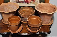 Five terra cotta pots of varied sizes are shown on a wooden surface with an apple basket painting in the background. The pots include two large scalloped rim pots, one medium pot, and two smaller pots each with matching trays. Visible wear and aging residue noted on pots.
