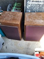 Two rectangular vintage speakers with wood veneer finish and maroon fabric grilles shown side by side on a floor with some storage items around.