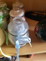 Three glass bottles of bath salts in pale blue, cream, and pink, each with a ribbon tied around the neck and sealed with glass lids.