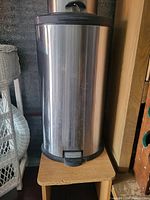 Full view of a cylindrical stainless steel garbage bin with black lid and foot pedal, placed on a wooden surface indoors.