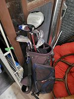 Photo showing a golf bag containing several golf clubs including a Ping G25 driver and multiple irons with steel shafts, bag has shoulder strap and front zippered pockets.