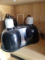 Front view of black dual-head heat lamp with white bulb sockets and central handle placed on wooden shelf