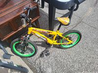 Yellow kids bike with green rims, black seat, and black handlebars. Bike positioned outdoors on concrete near some furniture and other items.