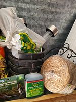 Photo shows close up of various outdoor gardening items including retractable plant hangers in packaging, green liquid plant food bottle, stacked black cast iron plant holders, brown coiled rope mat, and parts of black metal chair frames.