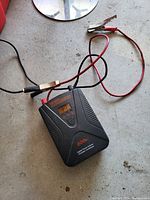 Power inverter placed on concrete floor showing front and top view including clip cables and digital display.