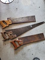 Top-down photo of three hand saws on a concrete surface showing wooden handles and rusted blades.