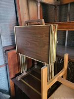 Multiple folding tables stacked together showing dark wood grain laminate tops with brass-colored frame edges and brown wood legs with rectangular base supports.