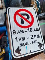 Photo of a rectangular metal No Parking sign showing the restricted parking hours: 9 AM - 10 AM and 1 PM - 2 PM Monday to Friday. The sign has double arrows pointing left and right and a red no parking symbol.