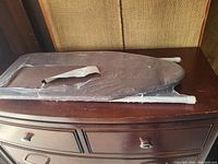 Photo of small ironing board folded with grey padded top and white metal legs on wooden surface