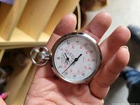 Close up photo of Foxlux brand mechanical stopwatch held in hand showing round chrome case, white dial with black and red markings and crown at top.