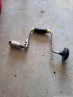 Photo of a hand crank drill with a black and yellow handle and a turning knob on the right side, lying on a concrete floor.