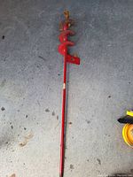 Photo showing a handheld ice auger with a long red metal shaft and a spiral blade designed for drilling ice.