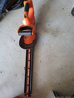 Black & Decker orange and black corded electric hedge trimmer with protective blade cover lying on a concrete floor.