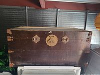 Front view of the large wooden trunk highlighting the metal handles, corner brackets, and central ornate lock plate on the dark stained wood surface.