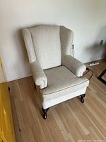 Light beige upholstered wing chair with dark wooden cabriole legs on a wooden floor next to a yellow cabinet.