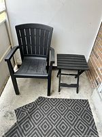 One black plastic patio chair and one black square slatted side table on a balcony floor with a patterned black and gray mat.