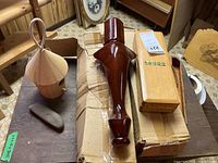 Four items displayed on wooden table: birdhouse, smooth wooden object, large polished turned wood table leg, rectangular artists box with label