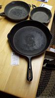 Three cast iron frying pans shown together on a wooden table: one with a long handle, two without handles but with loop grips.