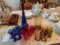Two decanters (blue with gold stripes and amber) with matching sets of glasses and additional unmatched glasses grouped on a table.