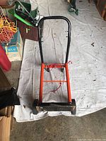 Top view of orange metal dolly with black handle, two wheels, and securing straps on white tarp in garage.