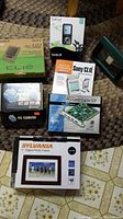 Overview of the lot items placed on a carpet: Sony Clie box, SanDisk Sansa e260 box, Sony Clie second edition book, PC camera, Nexxtech USB PCI card box, 4x6 wood photo frame, Sylvania 7 inch digital photo frame box
