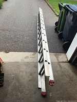 Full length view of aluminum extension ladder lying on garage floor extending out to pavement, showing ladder ends with red caps and overall structure.