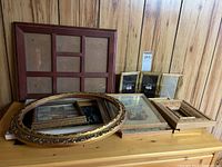 Overview of assorted frames stacked on a wood surface against a wood paneled wall, featuring oval ornate gold frame, multi-section frame, and several rectangular frames with prints