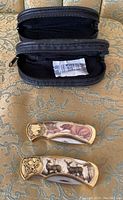 Two black zippered cases open, showing space for pocket blades, with two pocket knives laid out. Both knives show wildlife scene handles.