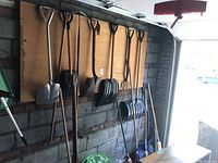 Wide view of tool rack in garage wall showing multiple garden tools hanging including shovels, snow shovels, rakes, brooms, pitch fork, and crow bar