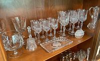 Photo showing one shelf with various clear glassware including stemmed wine glasses, liqueur glasses, a large snifter, a glass bell, and rectangular butter plate.