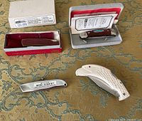 Four mini pocket knives shown on a patterned cloth, including two with boxes and papers